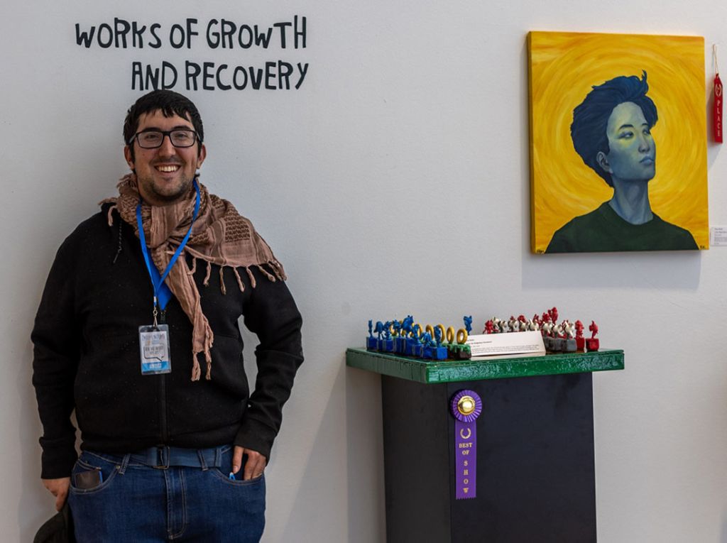 Winner of the Momentum Arts exhibition Works of Growth and Recovery standing next to his chess board which has a Best of Show ribbon